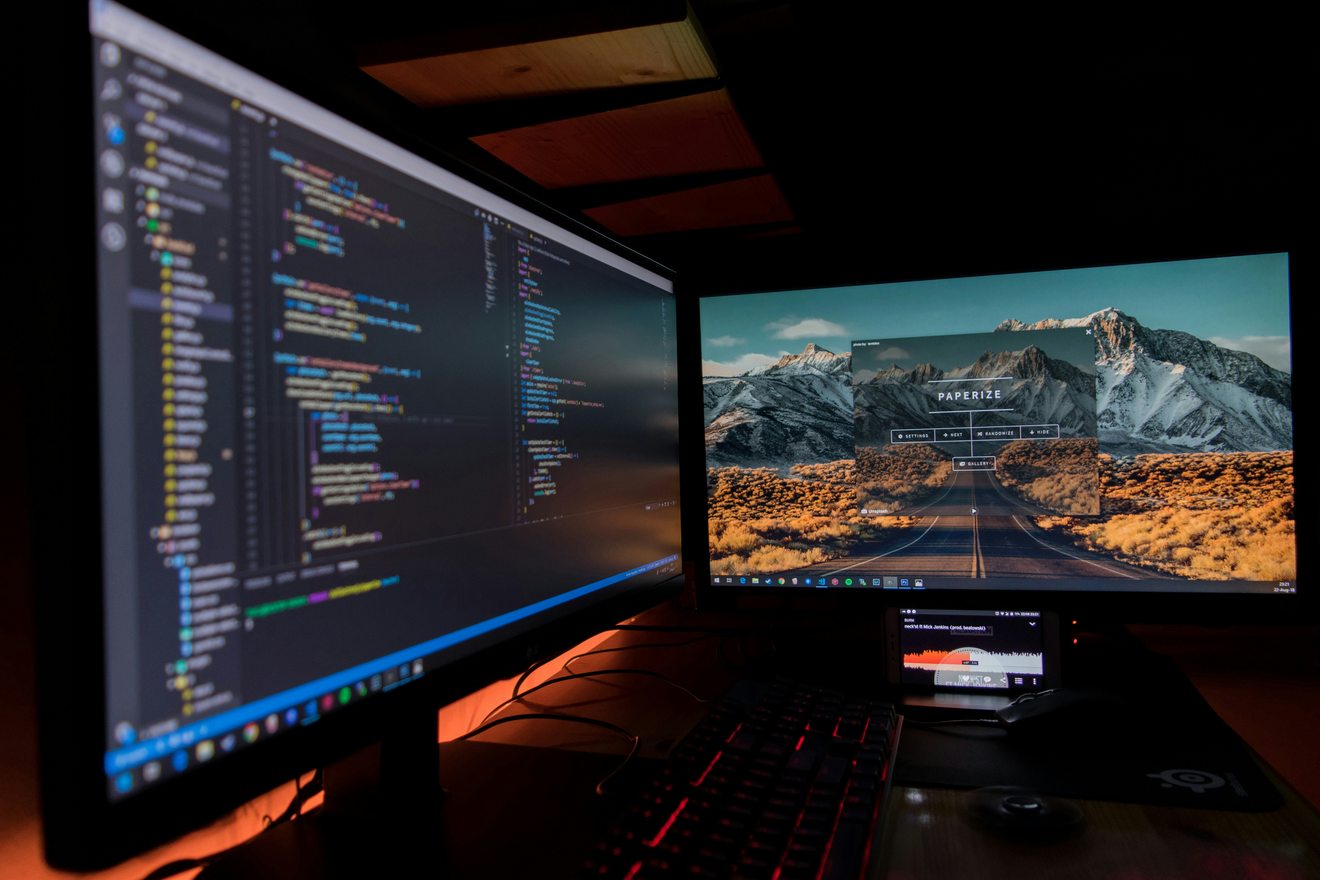 Two desktop monitors in a darkened room, one showing lines of code and one showing a design application