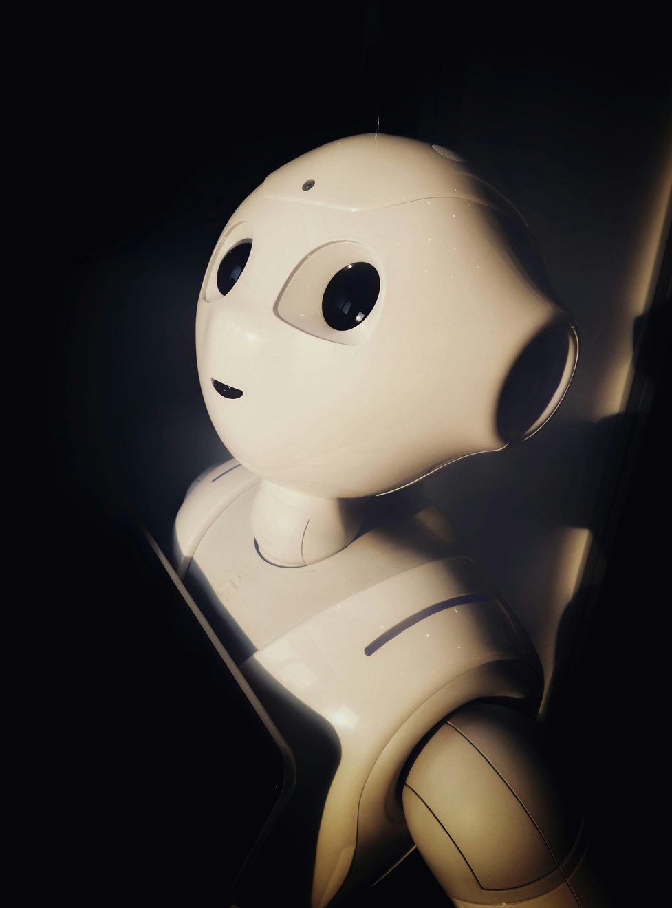 A white humanoid robot with large dark eyes, lit from one side against a black background