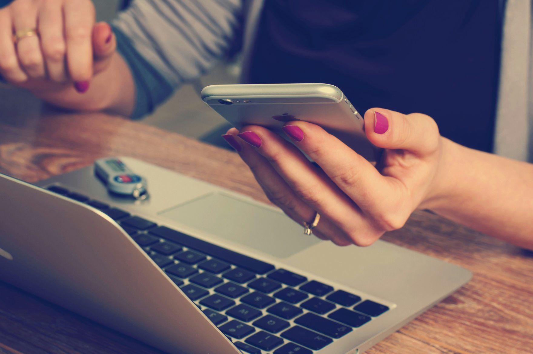 Two people at a wooden table, one holding a smartphone above an open laptop