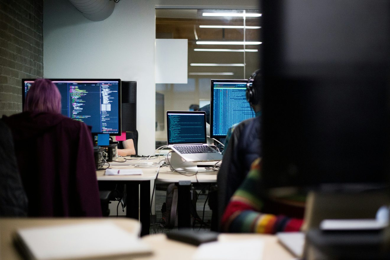 An open laptop and notebook on a shared desk in an office where two people are working