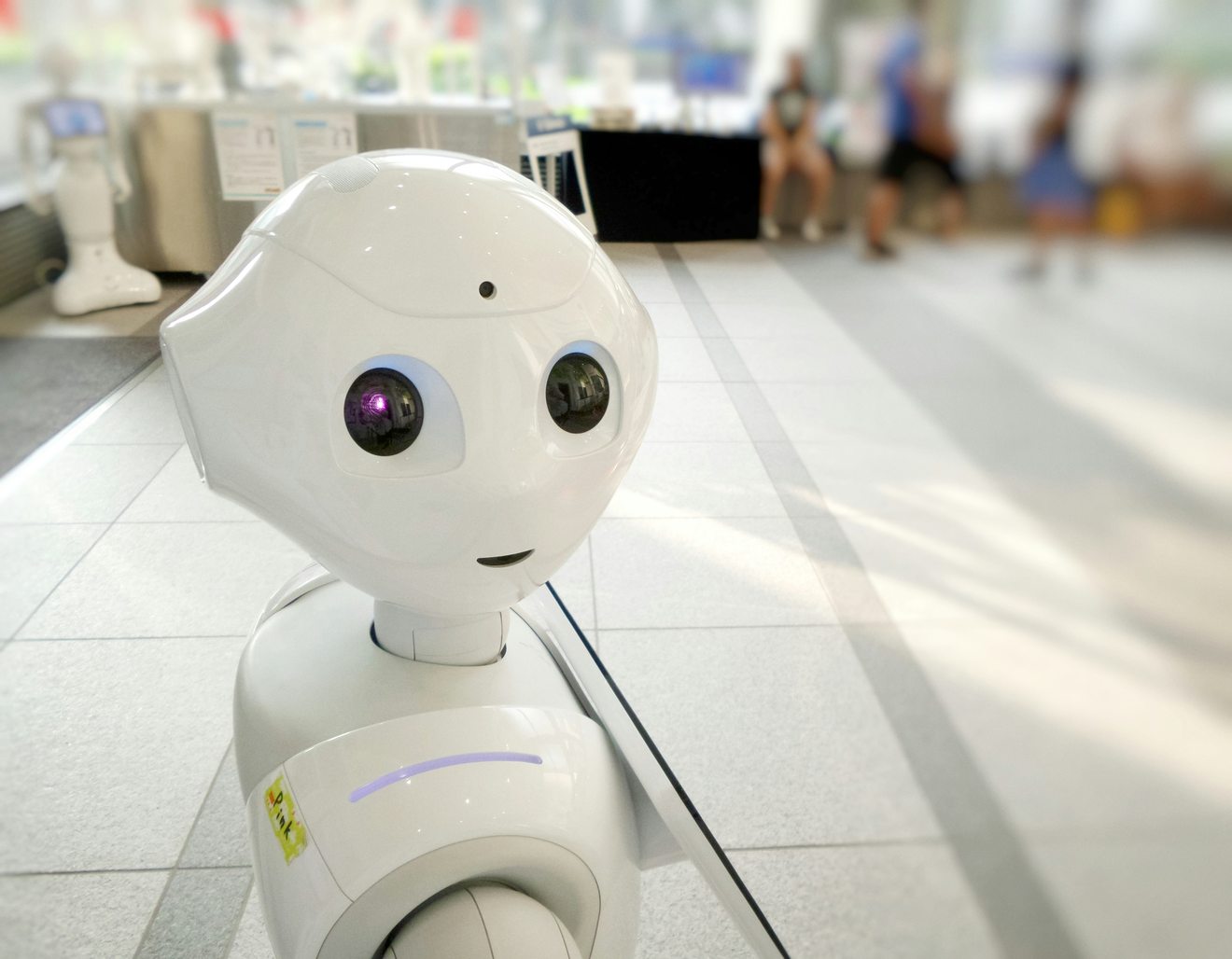 A white humanoid service robot on a tiled concourse with people seated in the blurred background