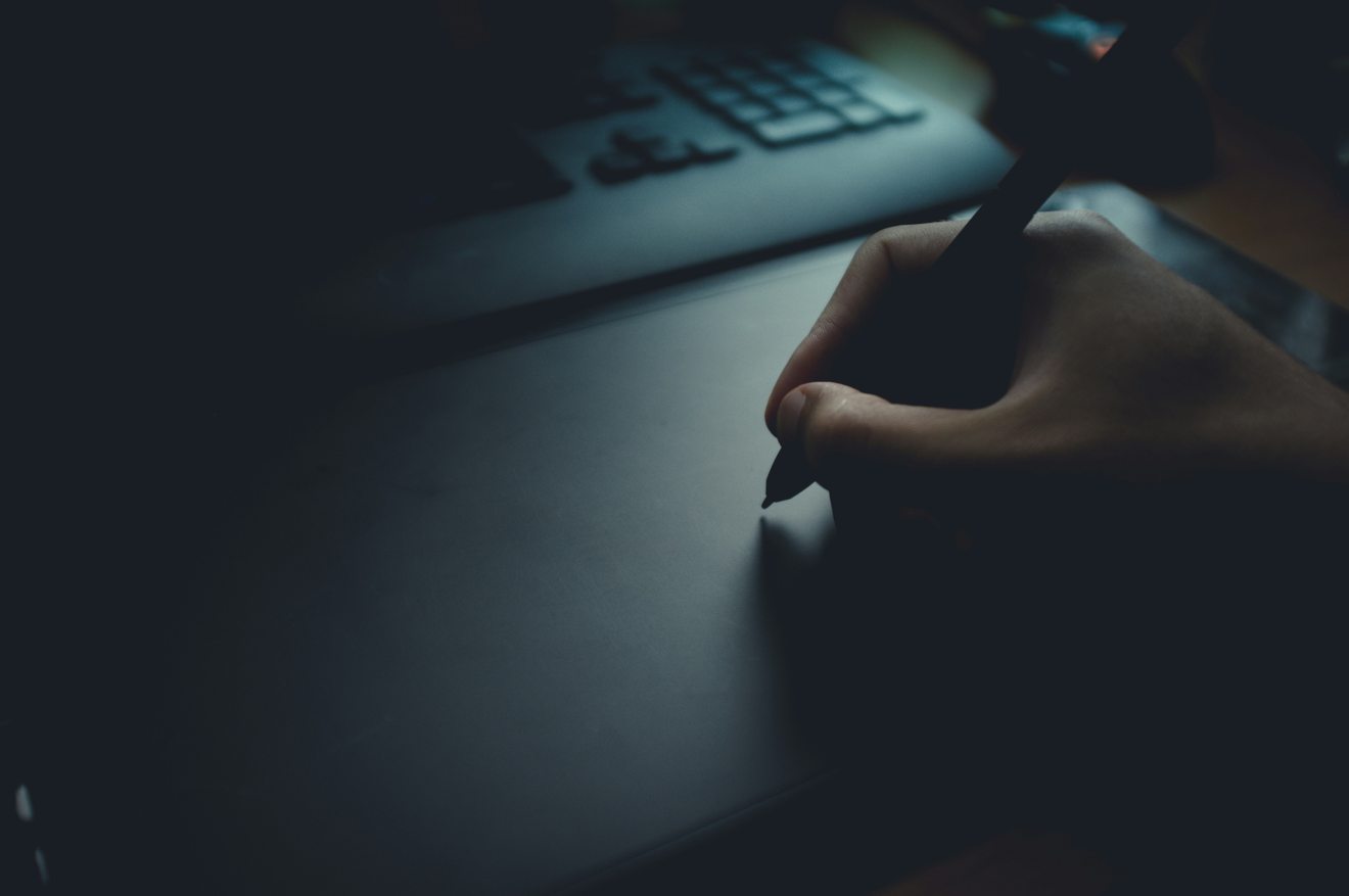 A stylus pressed to the surface of a drawing tablet in low light, with a keyboard behind it